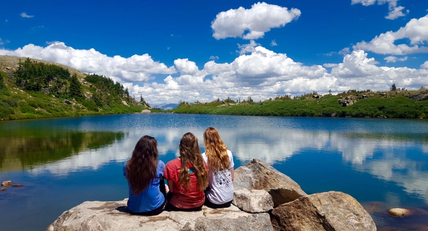 Three people sit on a rock near a body of water. They sky is reflected in the still water. 